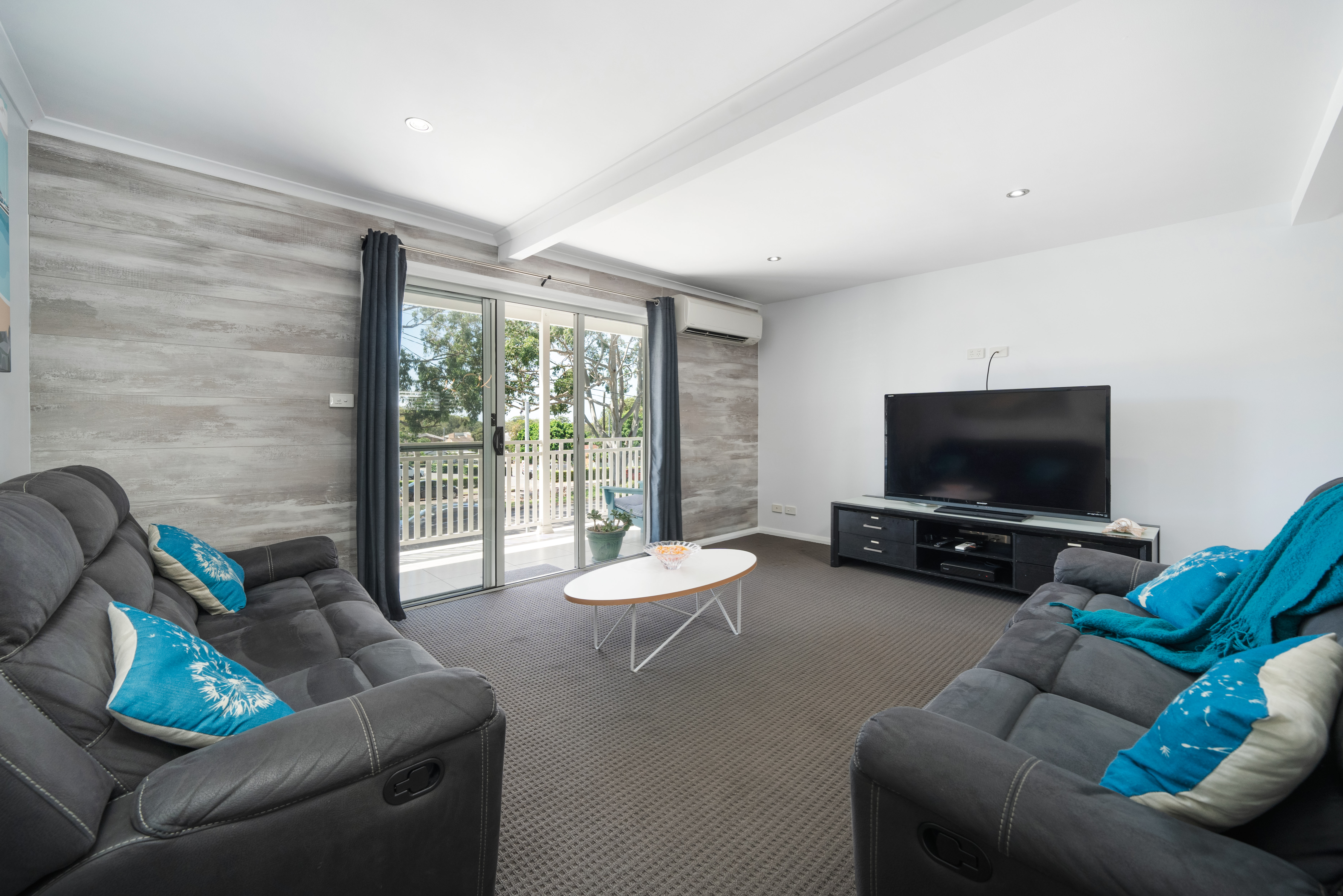 A photo of the main living room in the Beach and Bay Holiday House in Nelson Bay, which features a large flat screen TV, comfortable recliner lounges, air conditioning and a balcony.
